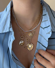 A person wearing a blue striped shirt showcases layered gold necklaces featuring various pendants, including the Mod + Jo Astra Fluted Charm. Among the collection are a yin-yang, a sunburst, and a heart adorned with a bird and arrows, as well as a bezel-set crystal pendant. Some of the necklaces are 14k gold plated. The person is holding their shirt collar.