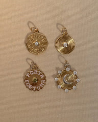 Four gold pendants on a beige textured background: a star hexagon, a sunburst with a bezel-set crystal, the Mod + Jo 14k gold plated Porta Fortuna disc, and the Astra Fluted Charm.