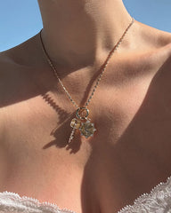 A person wearing the Mod + Jo Callum Tennis Charm Clip Necklace, featuring gold-plated key and flower-shaped charms, is shown against bare skin and a clear blue sky, with lace fabric visible at the bottom. The dainty chain necklace from Mod + Jo adds an elegant touch to this graceful scene.