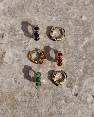 Three pairs of Mod + Jo Corynn Gemstone Huggies—each 18k gold plated with blue, green, or natural red agate stones—plus matching studs, beautifully displayed on a textured beige surface.