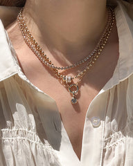 A person wearing a cream pleated blouse and three layered Mod + Jo Darcy Paperclip Enhancer Chains in 14k gold plate, featuring beads, rhinestones, and a circular pendant with a charm.