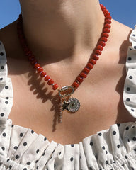A close-up of a person wearing the Mod + Jo Demi Tennis Charm—a red beaded necklace adorned with silver charms, including a “FORTUNA” coin, a key, and a sparkling CZ—over a white ruffled top with black polka dots.