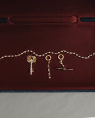 The Mod + Jo Demi Tennis Charm set showcases a dazzling 14k gold-plated chain and matching earrings, both designed with circular tops and delicate dangling accents featuring a sparkling CZ charm, beautifully presented on a deep red velvet backdrop.