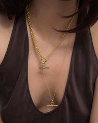 A person wearing a dark, deep V-neck top showcases two layered Mod + Jo Elliot Toggle Charm necklaces in 14k gold, each featuring a horizontal bar pendant and distinct chain styles, set against their bare upper chest.