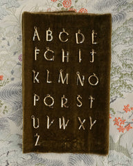 A set of Eloise Letter Charms by Mod + Jo, gold plated and arranged in four rows on a dark velvet board, displayed over patterned fabric with floral and wave designs.
