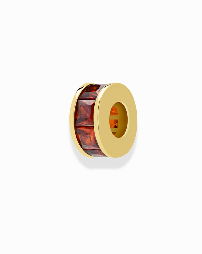 The Mod + Jo Este Birthstone Charm Spacer is a gold spacer bead with a hollow center and a row of rectangular red gemstones, ideal for personalizing your jewelry collection. Displayed on a white background.