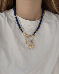 A person wearing a white shirt with three layered necklaces: one gold plated letter charm necklace, one with dark blue beads, and the Mod + Jo Este Birthstone Charm Spacer—a chic blend of personalized accessories.