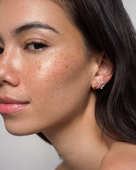 A woman with smooth, freckled skin and long dark hair wears the Gabby Marquise Dangle Studs by Mod + Jo—two small, gold-plated earrings delicately shaped like leaves. She gazes softly at the camera against a plain background.