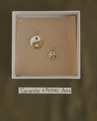 Two Mod + Jo Grande Ava Yin Yang charms, one large and one small 14k gold pendant, are displayed in a box on brown paper. Below the box is a label that reads "Grande + Petite Ava" on a green fabric background.