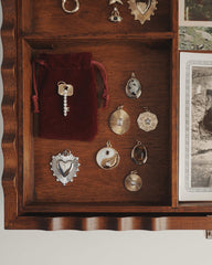 A wooden drawer holds an assortment of vintage charms and pendants, including the Grande Ava Yin Yang Charm from Mod + Jo, nestled among heart-shaped, spiral, and sunburst designs. A small red velvet pouch and a glimmering mother-of-pearl necklace rest beside the trinkets, while black-and-white photos peek in from the right.