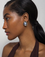 A woman with smooth brown skin and long dark hair in a low ponytail wears a sleeveless brown top and Mod + Jo's Hunter Statement Studs—large oval silver earrings set with blue chalcedony—looking left against a light background.