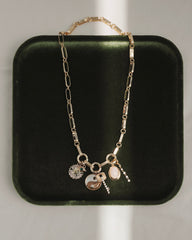 The Jagger Charm Clip Necklace by Mod + Jo, crafted in 14k gold and featuring four unique charms—a yin-yang, a flower, an oval stone, and a petite hanging pendant—is elegantly displayed on a dark green velvet tray with sunlight creating gentle shadows.