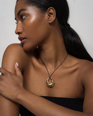 A woman with dark skin and long, dark hair in a low ponytail poses in profile, wearing a black strapless top and the Mod + Jo Kira Cord Necklace, featuring an adjustable cord and gold pendant, her arm gently resting across her chest.