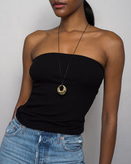 A woman in a black strapless top and blue jeans poses against a plain background, accessorized with the Mod + Jo Kira Cord Necklace, which features an adjustable cord and a striking gold pendant.