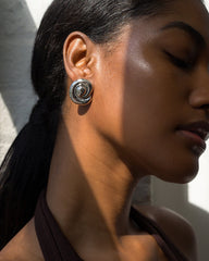 A woman with smooth, glowing skin wears Mod + Jo Milley Spiral Statement Studs and a brown halter top, tilting her head slightly in soft, natural light with gentle shadows across her face.