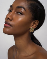 A woman with glowing skin and long dark hair wears Mod + Jo’s Milley Spiral Statement Studs in 14k gold. She gazes confidently at the camera, showcasing these elevated everyday earrings against a plain background.