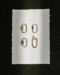 Four metallic Pip Charm Enhancers by Mod + Jo—three in radiant 14k gold and one in lustrous silver—are arranged in a grid on a white, rough-edged sheet of paper set against a dark background. A beam of light falls vertically across the arrangement, showcasing each enhancer like an elegant, modern charm accessory.