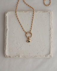 The Nyx Evil Eye Charm necklace by Mod + Jo features a round beaded gold chain and an eye-shaped pendant set with a green gemstone, artfully displayed on a textured white square tile—an elegantly crafted piece of protective jewelry that exudes the subtle allure of the evil eye.