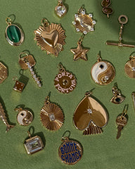 Mod + Jo gold and jeweled charms—including hearts, stars, yin-yangs, a protective pendant, and the Nyx Evil Eye Charm—are displayed on a green textured fabric background.