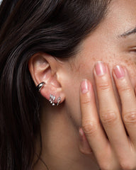 A close-up of a person’s ear adorned with three earrings: a silver hoop at the top and two gemstone studs in the lobe, one of which is Mod + Jo’s Gabby Marquise Dangle Studs that catch the light beautifully. The person’s neatly manicured hand rests gently against their face.
