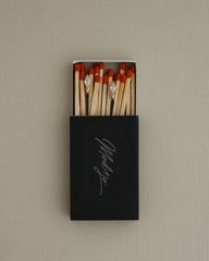 A black matchbox with “Marlsto” written on it, filled with matches, displays two matches adorned with sparkling Phoebe Kite Huggies by Mod + Jo—each shining like dazzling cubic zirconia earrings—set against a neutral textured background.
