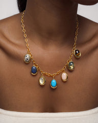 A close-up of a person wearing Mod + Jo's Rory Gemstone Charm necklace, featuring seven vibrant gemstone pendants—including a striking lapis lazuli—set in 14k gold over brass. Paired with a white off-shoulder top, the jewelry adds an elegant touch to the look.
