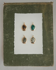 Four Rory Gemstone Charms by Mod + Jo, each featuring a unique oval gemstone with 14k gold over brass edges, are arranged on a sheet of white paper centered against a textured olive green square background. Each charm showcases a different stone—including the striking Dalmatian Jasper—and is thoughtfully crafted for an elegant touch.