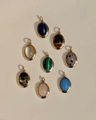 Eight exquisite Rory Gemstone Charms by Mod + Jo, each framed in 14k gold, are elegantly arranged on a light surface. Every charm showcases a distinct gemstone hue—serene blue, vibrant green, mysterious black, luminous white, gentle pink Rose Quartz symbolizing self-love, and uniquely patterned multicolored stones.