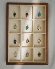 A wooden display case featuring twelve compartments arranged in a 3x4 grid, each showcasing a Mod + Jo Rory Gemstone Charm—a round pendant with a unique colored stone such as exquisite lapis lazuli—beautifully illuminated by natural sunlight.