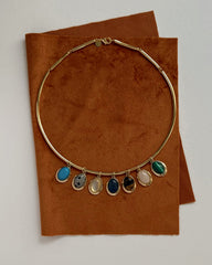 The Mod + Jo Rory Gemstone Charm necklace, featuring seven colorful gemstone pendants and inspired by lapis lazuli jewelry, is displayed on brown suede fabric against a white background.