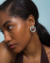 A woman with smooth, dark skin and long, dark hair pulled back wears the Mod + Jo Sabine Statement Earrings—bold double-loop shapes—as she looks to the side against a light blue background.