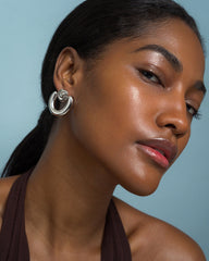 A woman with smooth, glowing skin models the Sabine Statement Earrings by Mod + Jo. She wears a brown halter top and has dark hair pulled back, posing confidently against a light blue background.