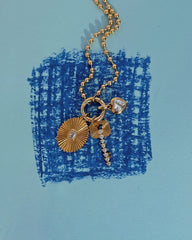 The Soren Ball Charm Clip Necklace by Mod + Jo, featuring a gold plated design with three charms—a sunburst disc, a rhinestone-accented key, and a heart-shaped gem—is displayed on a blue paper background with a textured blue square sketch.