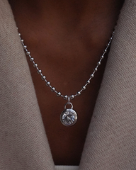 Silver ball chain necklace with a round sapphire bezel pendant on a person wearing a beige cardigan.