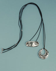 Two Suna Spiral Cord Necklaces by Mod + Jo rest on a light blue surface: one has a shiny double-spiral pendant, the other features a reflective crescent charm. Each adjustable piece blends unique style with comfort.