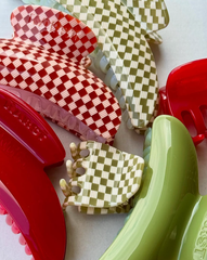 Close-up of several large SuperClip Hair Claw clips in red, green, and cream—some with a checkered pattern—overlapping and artfully arranged on a light surface.