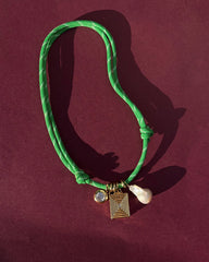 The Mod + Jo Tommy Cord Necklace features a green paracord and three charms—a round gem, a geometric gold pendant, and a white pearl—shown on a burgundy background.
