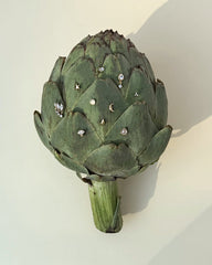 A green artichoke displays an assortment of small, delicate jewelry—gold earrings, rings, and Mod + Jo’s Uma Teardrop Studs—arranged on its leaves against a light beige background.