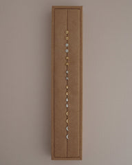 A brown rectangular jewelry display showcases the Mod + Jo Uma Teardrop Studs, along with other small stud earrings in gold and rhodium plated silver, featuring circular, star, teardrop, and crescent moon shapes. These high-shine earrings are arranged vertically against a neutral background.