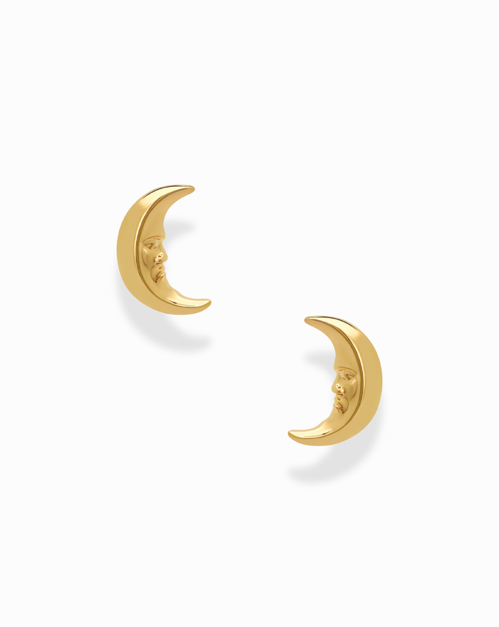 The Archie Moon Studs by Mod + Jo are a pair of gold plated earrings, each adorned with a delicately carved crescent moon face. Displayed on a white background, these whimsical studs make an enchanting addition to any celestial jewelry collection.