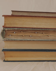 A stack of four old books lies horizontally, with Mod + Jo’s Archie Moon Studs inserted along the edges of the pages in the second book from the top. The background is neutral and plain.
