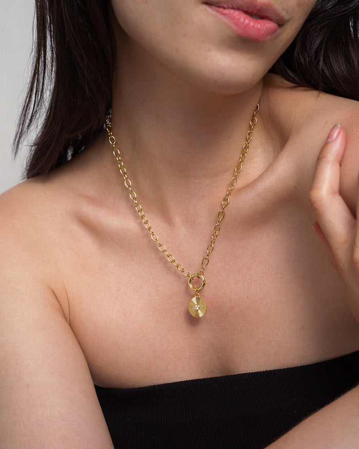 A woman wearing the Mod + Jo Astra Fluted Charm—a 14k gold plated chain necklace featuring a round, bezel-set crystal pendant—poses with her hand near her collarbone while dressed in a black, strapless top against a neutral background.