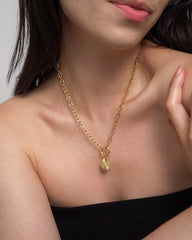 A woman wearing the Mod + Jo Astra Fluted Charm—a 14k gold plated chain necklace featuring a round, bezel-set crystal pendant—poses with her hand near her collarbone while dressed in a black, strapless top against a neutral background.