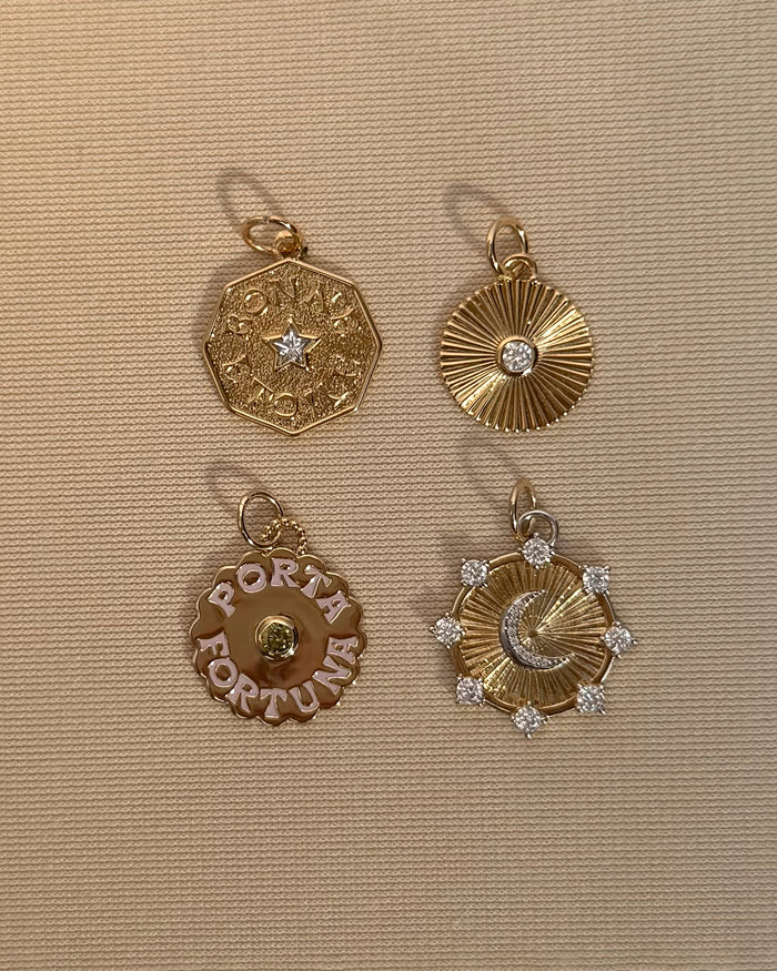 Four gold pendants on a beige textured background: a star hexagon, a sunburst with a bezel-set crystal, the Mod + Jo 14k gold plated Porta Fortuna disc, and the Astra Fluted Charm.