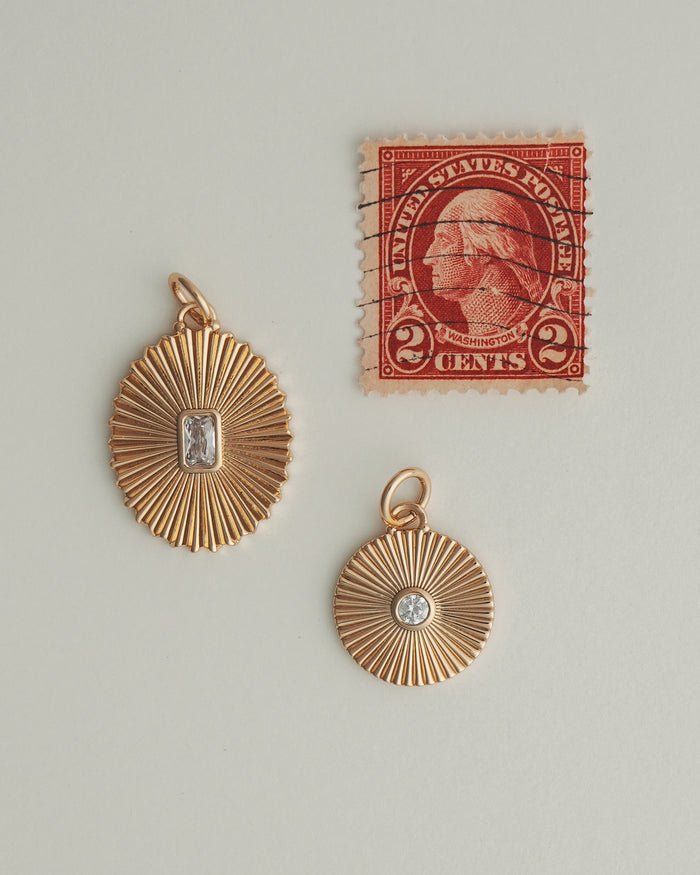 Two Mod + Jo Astra Fluted Charms, featuring bezel-set crystal center stones in a gold sunburst design, are displayed beside a vintage red two-cent US postage stamp with a profile portrait.