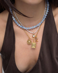 A woman wears a brown top and three layered Mod + Jo necklaces: one with blue beads, one gold chain, and one featuring playful gold coin jewelry—including a hexagon-shaped pendant, a square locket, an initial R charm, and the Bonne Etoile Charm for an added touch of luck.