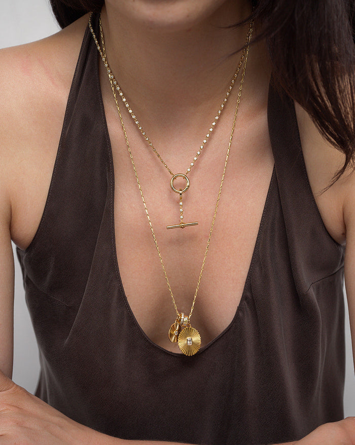 A woman wearing a deep brown top and two layered gold necklaces: a dainty chain necklace with a horizontal bar pendant, and the Mod + Jo Callum Tennis Charm Clip Necklace featuring a round, textured pendant with a small gold charm.