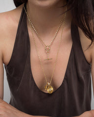 A woman wearing a deep brown top and two layered gold necklaces: a dainty chain necklace with a horizontal bar pendant, and the Mod + Jo Callum Tennis Charm Clip Necklace featuring a round, textured pendant with a small gold charm.