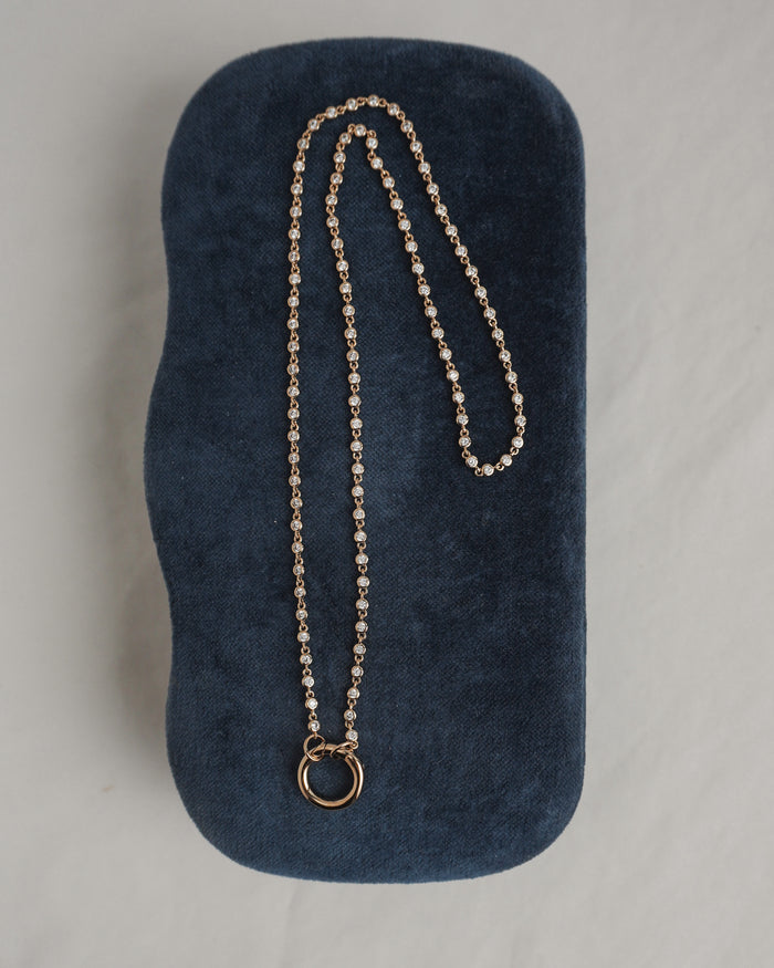 The Mod + Jo Callum Tennis Charm Clip Necklace, featuring a circular pendant and gold plating, is displayed atop a dark blue, velvet-textured case on a light surface.