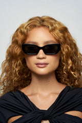 A woman with curly, light brown hair wears Elisa Johnson’s Cookie Sunglasses—black cat-eye frames with UV400 protection—and a black top with a twisted neckline detail. She faces forward against a plain, light background.
